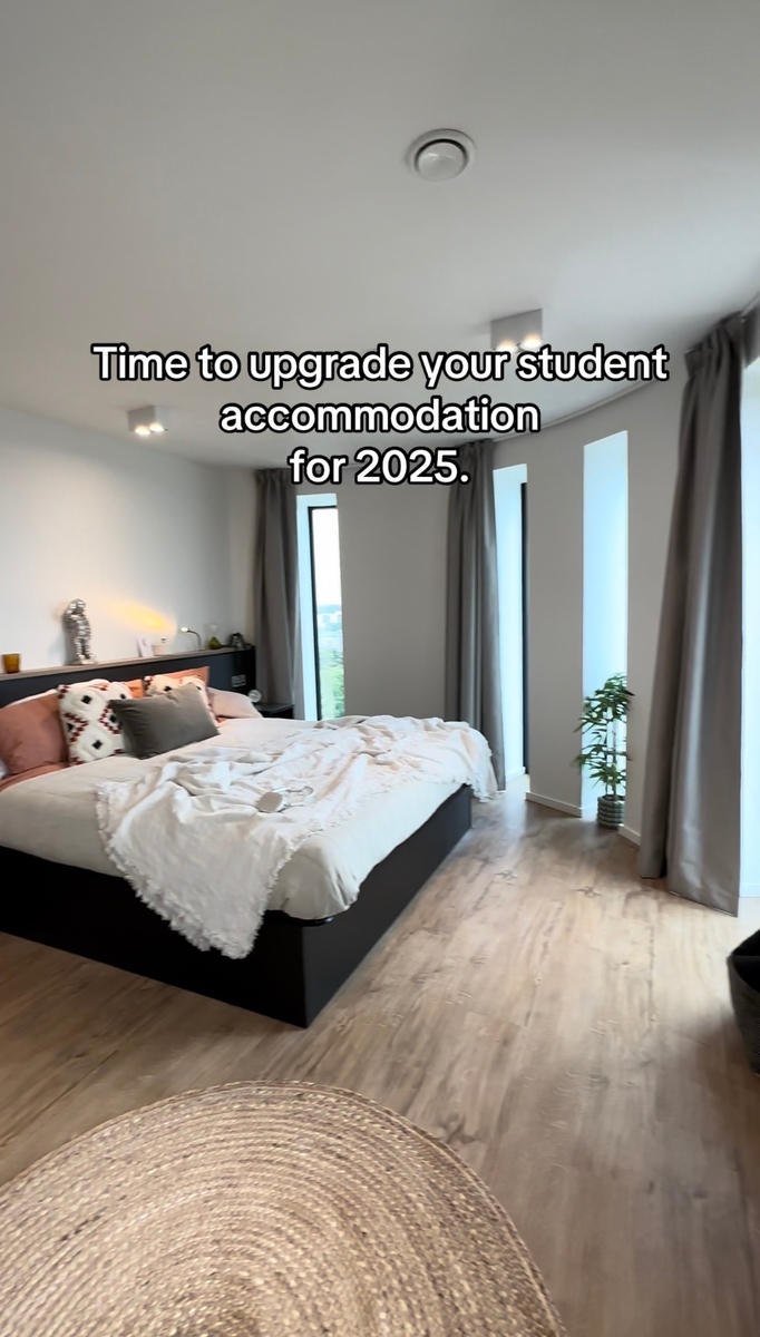 A modern bedroom with a large bed featuring white linens and decorative pillows. The room has wooden flooring, tall windows with gray curtains, and a potted plant. Text on the image reads, "Time to upgrade your student accommodation for 2025.